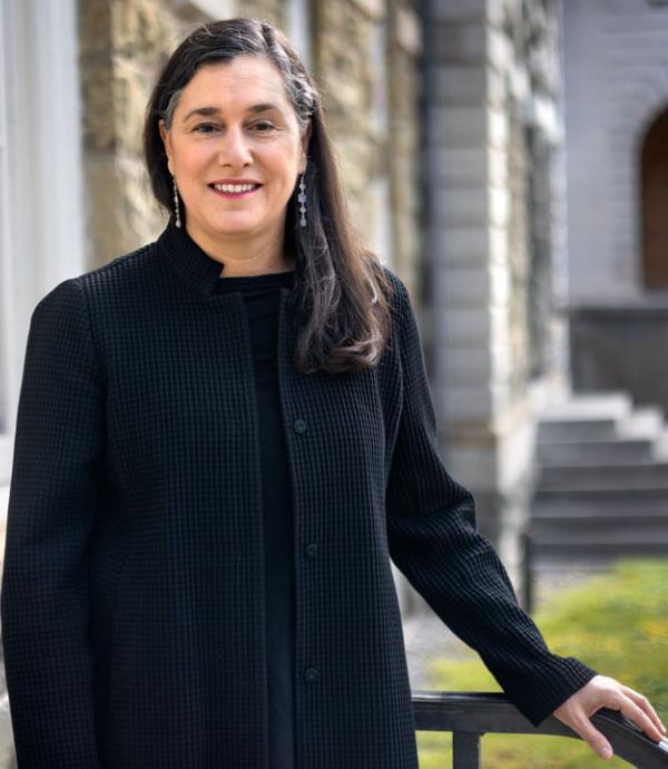 a person with long hair in a black coat smiling at the camera with one hand resting on a railing